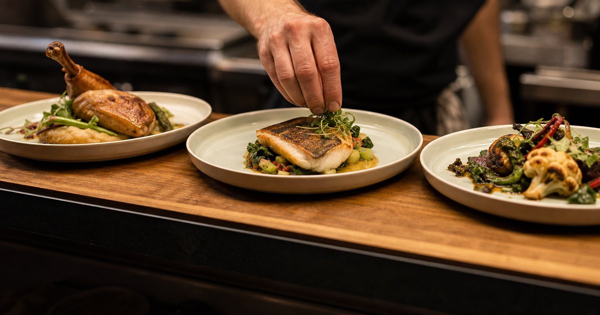 Three plated dinners side by side on a wooden restaurant pass, roasted chicken, seared fish, and a vegetable dish, a chef's hand adjusting a garnish, warm kitchen light