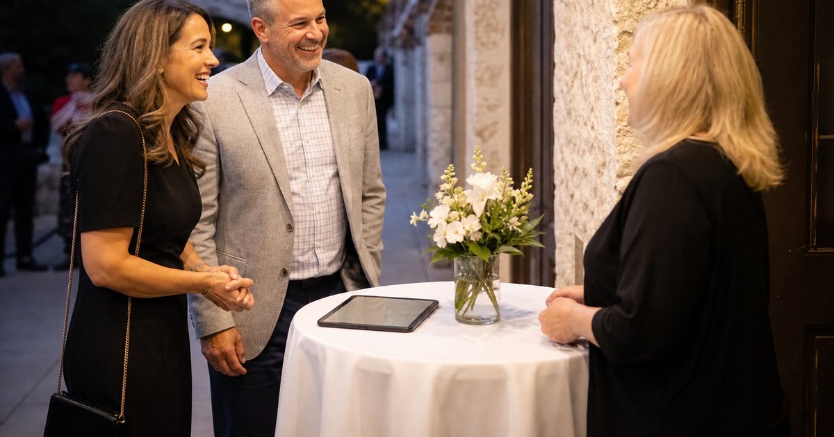 A woman at a small welcome table outside an event hall greeting a smiling couple, a vase of flowers on the table beside her, evening light