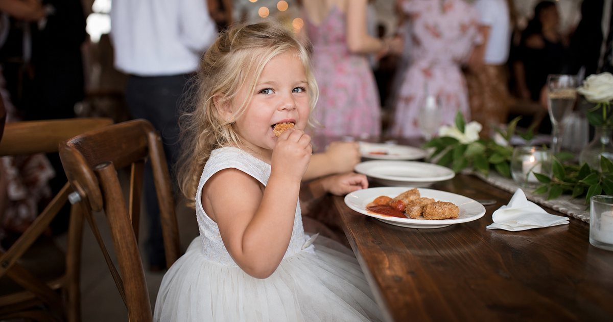 A small child in a party dress sitting at a decorated reception table eating chicken tenders from a plate, adults blurred in the background, string lights overhead