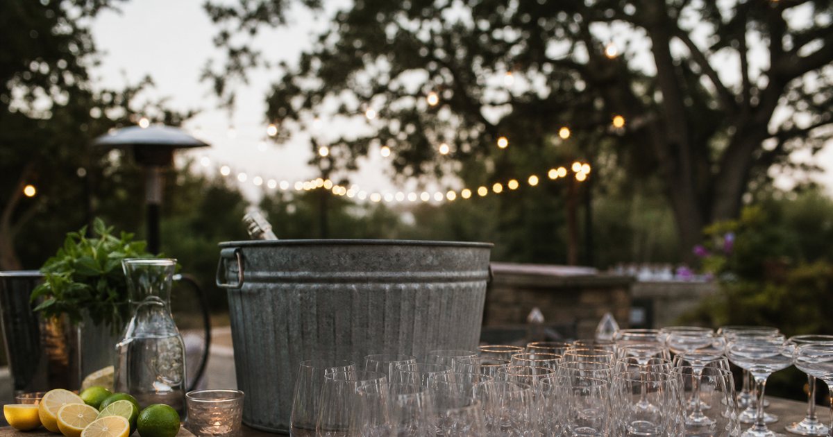 An outdoor evening reception bar on a rustic wooden table, rows of empty stemless wine glasses and coupe glasses, a galvanized ice bucket, halved lemons and limes on a cutting board, warm string lights overhead