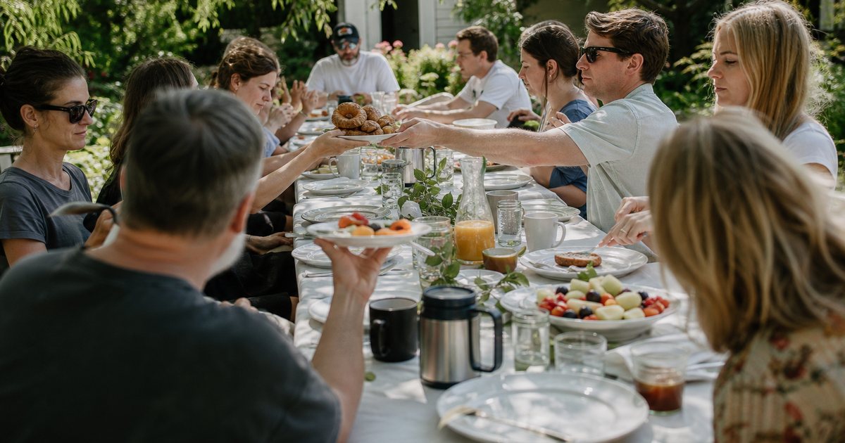 A long outdoor brunch table in a garden the morning after a wedding, guests in casual clothes passing plates of pastries and fruit, coffee carafes, dappled sunlight