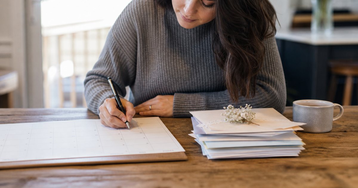 A woman at a kitchen table sealing a plain cream envelope, a neat stack of blank envelopes and a coffee mug beside her, soft morning window light